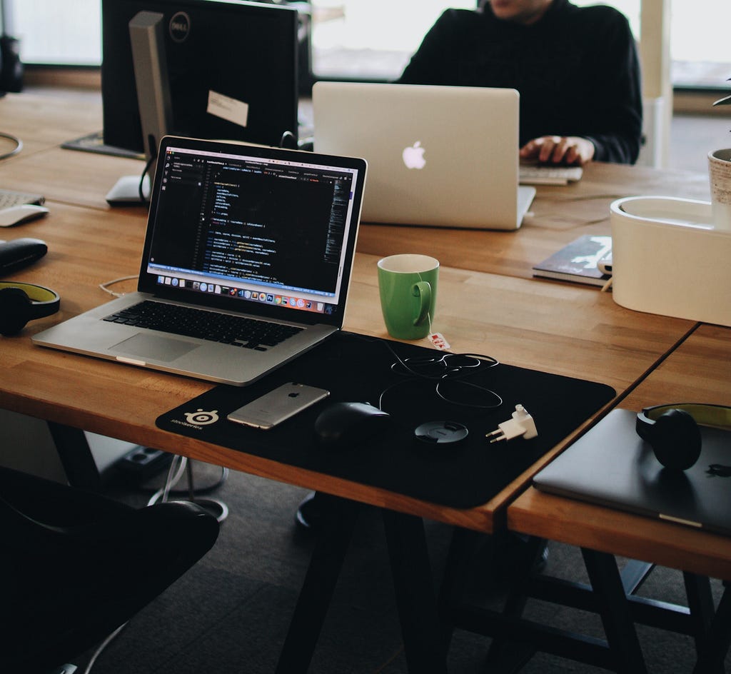 Laptop, coffee mug and iphone on a brown desk in front of a man in black sweater also on laptop.