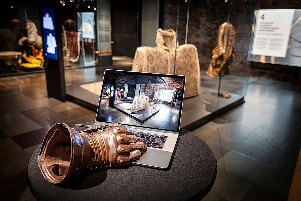 In the background a museum space with Swedish royal wedding clothes from the 1800s. In the foreground an armour glove on a laptop computer.