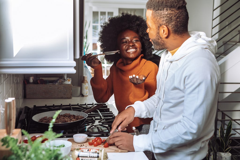 Foto colorida apresenta um casal negro cozinhando. A mulher negra está vestida com um moletom na cor laranja, segurando uma colher de pau enquanto sorri, o homem, também negro, está vestindo um casaco branco, enquanto pica alguns tomates e olha para a mulher, eles estão em uma cozinha perto do fogão.