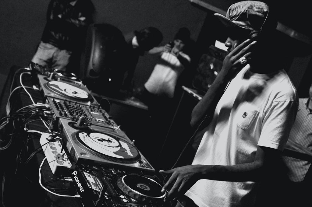 A man in a cap and white t-shirt stands behind a turntable set-up. His left hand is touching a turntable controller and his right hand is touching his headphones. There are people standing in the background. This is a black and white image.