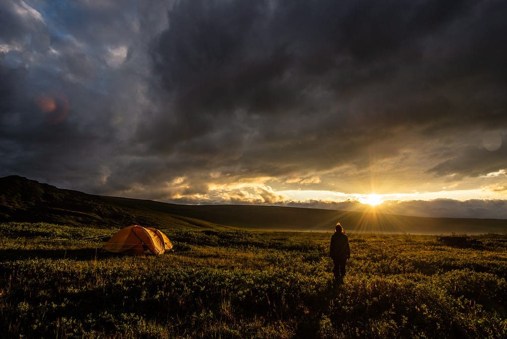 A person looks over fields and mountains into the setting sun with dark storm clouds and orange tents.