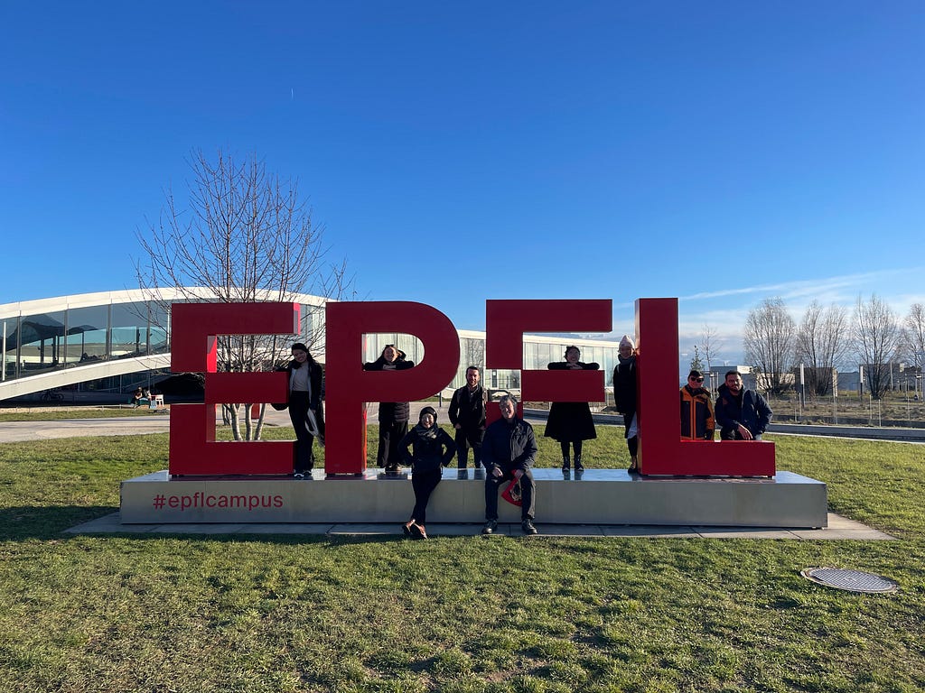 People stand and sit on a raised concrete platform with the letters “EPFL” carved into red statues; the grass is green in the foreground and blue skies in the background.