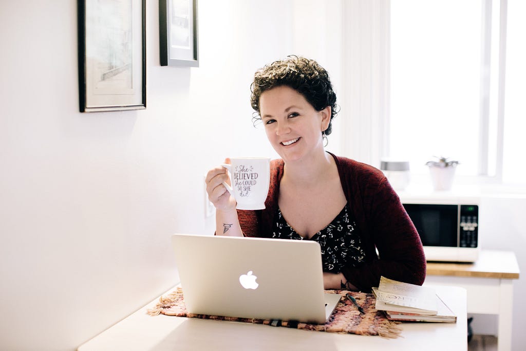 Renee sits smiling behind a laptop, holding a cup of coffee that says “she believed she could, so she did”