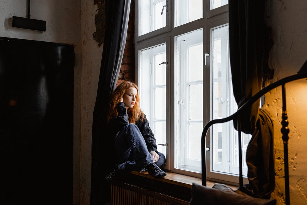 Woman sitting next to a large window in a dim room.