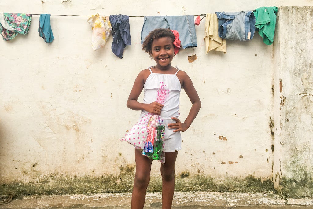 A Afro-Brazilian girl stands in front of a off-white wall, holding a bag of hygiene supplies.