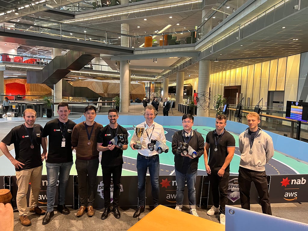 Eight people standing side-by-side in front of the miniature-sized racing track. One person is holding the trophy and one of the vehicles used. Two other people are holding the vehicles used. Seven of the eight people are wearing a medal around their neck.