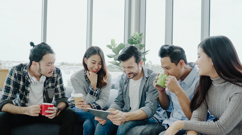 Group of young peers sitting on a couch drinking coffee by an office window looking at an iPad together.