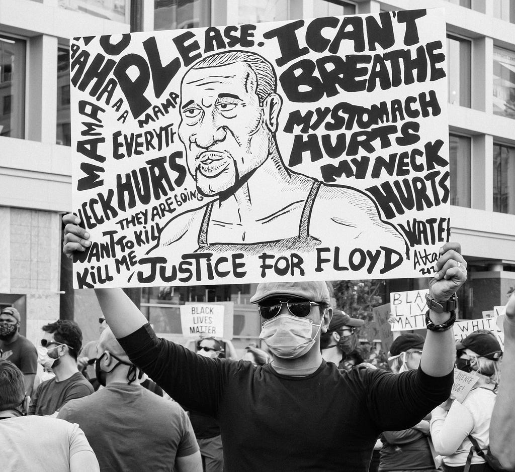 Greyscale photo of a man holding up a sign at the Black Lives Matter protest in Washington, DC on June 6, 2020. The sign that the man is holding is a drawing of George Floyd and at the bottom states “Justice for Floyd”. Other words and statements surround the poster including: “Please”, “I can’t breathe”, “My stomach hurts”, “My neck hurts”, “Water”, “No”, “Everything hurts”, “They are going to kill me”, “They want to kill me”.
