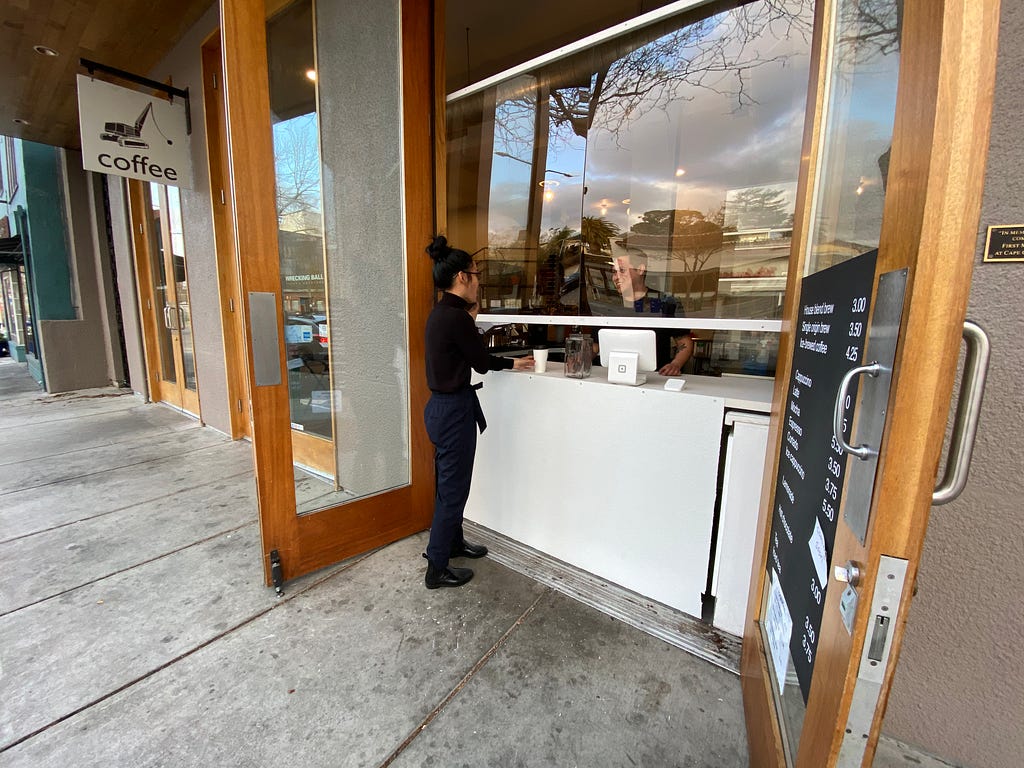 Open double-doors reveals a white counter and glass partition as a person is served coffee from a barista.