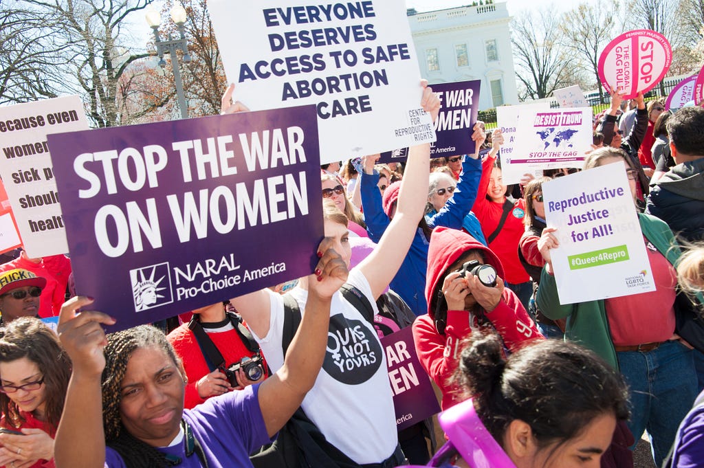 People advocating for reproductive rights/reproductive health. Signs stating “everyone deserves access to safe abortion care,” “stop the war on women,” and “reproductive justice for all.”