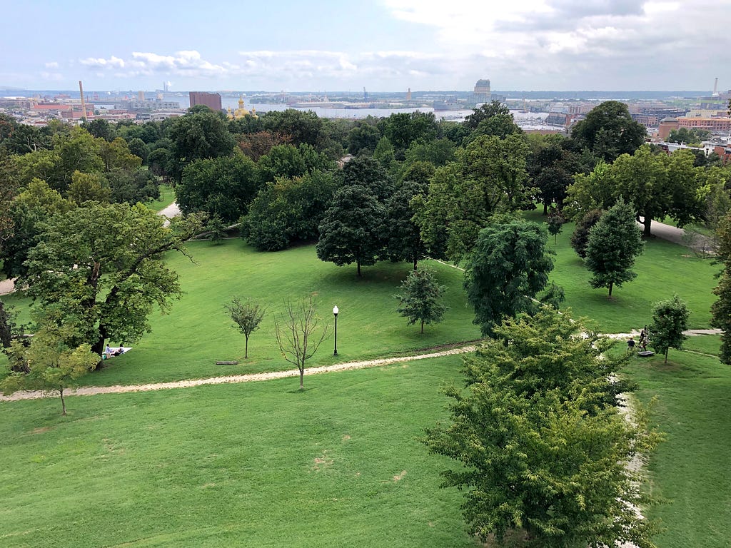 Photo of Patterson Park, in Baltimore, taken from high up in an observation building near the top of the park. The city is visible in the distance. The park is green. It is summer.