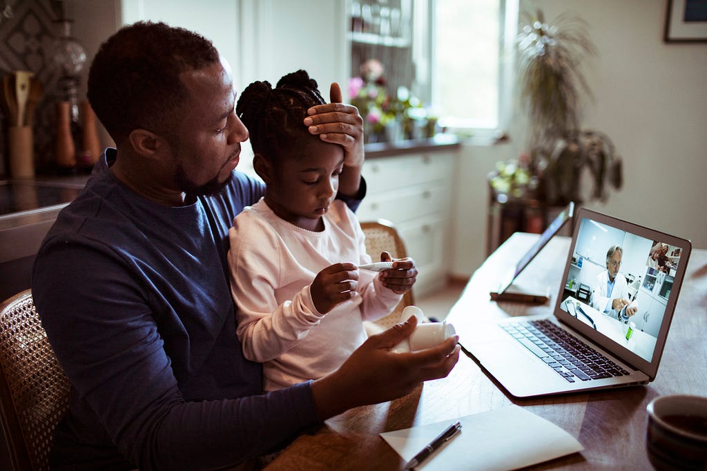 Father checking daughter’s forehead for fever during a telehealth appointment. Photo by Geber86/Getty Images