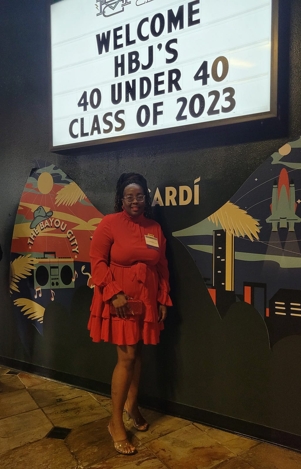 Kara standing in front of a sign that says Welcome HBJ’s 40 under 40 Class of 2023 writing in black letters. She is wearing a long-sleve red dress that has a high neck and a layers flowy bottom. The dress also has a belt that is tied in the front. Kara hair is in braids and in a high ponytail. The wall behind Kara has a painting with butterfly wings that says The Bayou City. The wings have pictures of a radio, music box, birds, the sun, a city skyline and rocket.