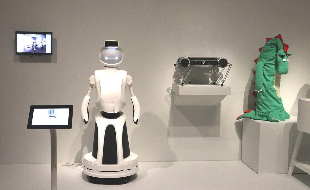 Three different robots in museum exhibit, one is human-shaped, one is a flat block with legs, last is wearing a dino costume