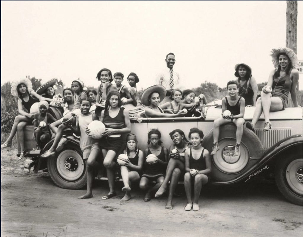 A black-and-white photograph of a large group of Black adults and children gathered around and seated on a car during a picnic, smiling and holding food outdoors.
