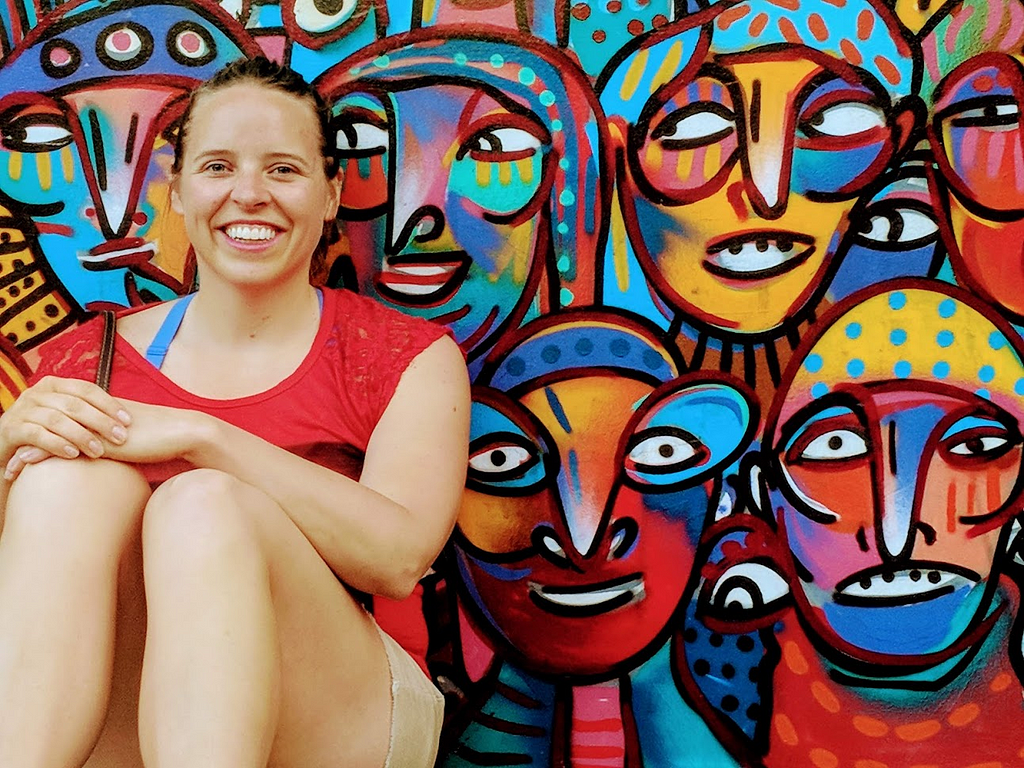 A woman smiles in front of a painted mural of many colorful faces