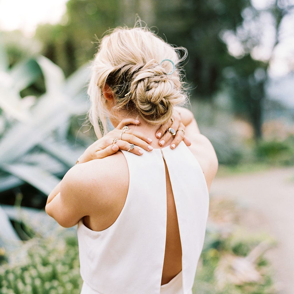 Image of blonde, white woman with a white dress, holding her neck with both hands. Greenery in the background, blurred.