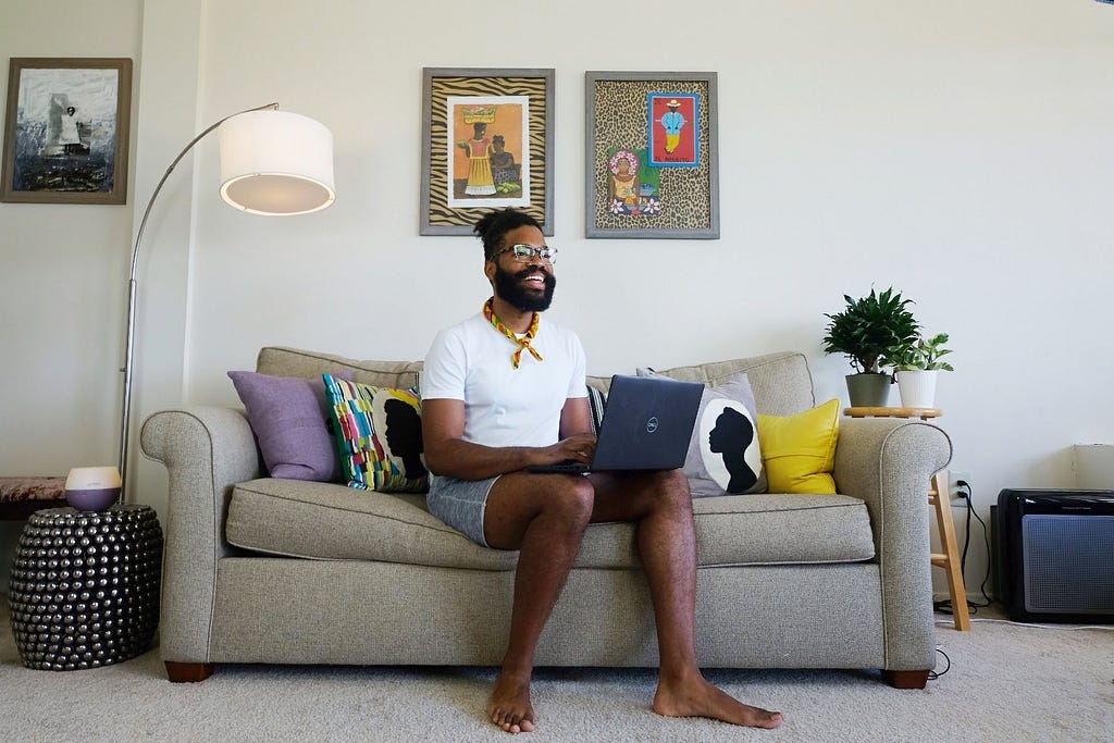 Black man sits on couch smiling with laptop.