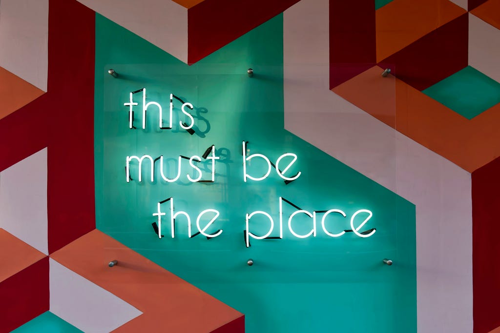 A neon sign on a vibrantly painted wall reads “this must be the place”, presumably situated in a workspace designed to offer a good work-life balance [Credit: Tim Mossholder on Unsplash].