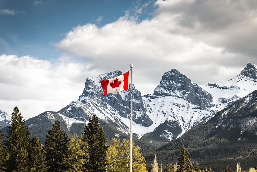 Canada Canmore, Alberta. Canadian Flag with Three Sisters in the Background. Photo and text by Igor Kyryliuk on Unsplash