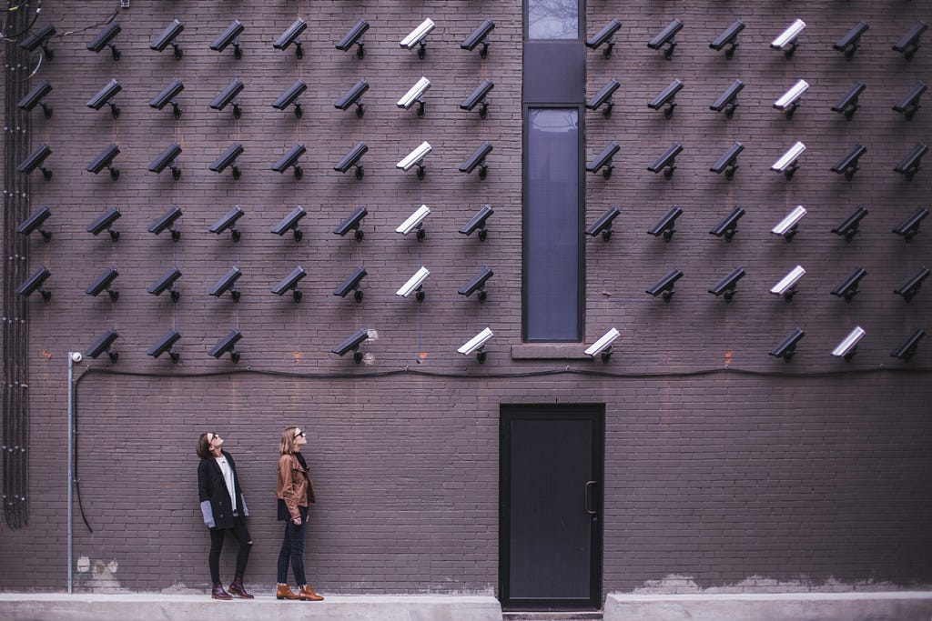 Two women standing next to a wall, looking up at the wall covered in surveillance cameras all pointing at them.