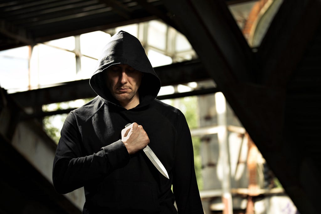 Image of a man wearing a black hoodie, standing in an industrial setting with a serious expression. He holds a large knife pointed downward in his right hand, creating a tense and ominous atmosphere.