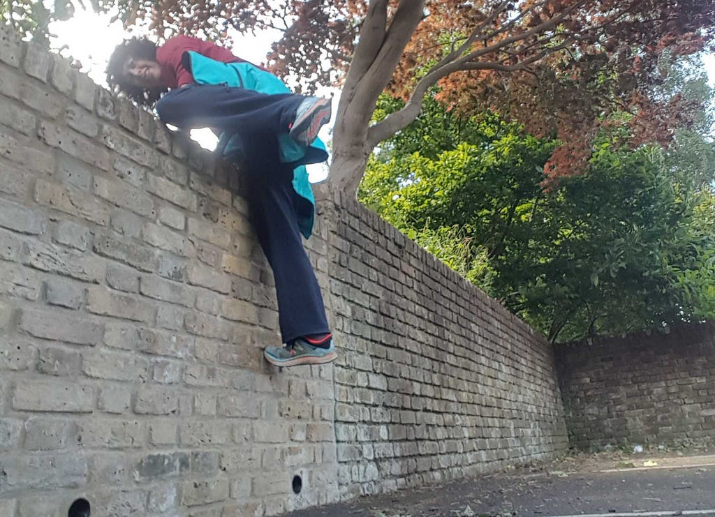 A woman in a tracksuit climbing onto a chest-high brick wall under a tree