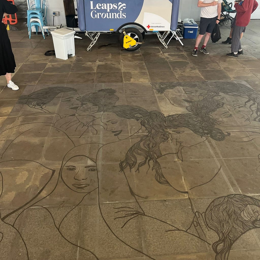 A mural adorns the floor in front of the Leaps & Grounds coffee cart. It depicts a diverse group of women and young girls.