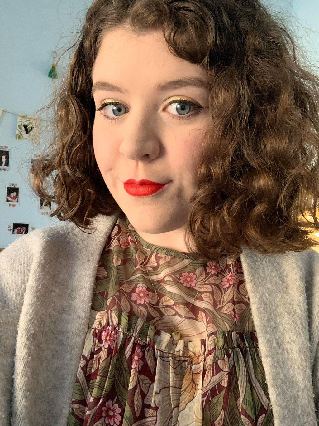 Anna is a white non-binary person with shoulder-length brown hair. She wears lipstick, a patterned blouse and grey cardigan.