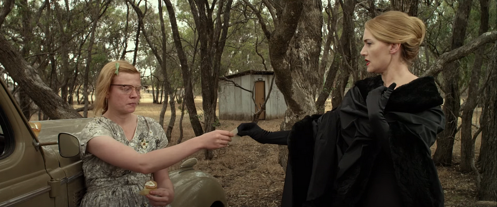 A screencap from The Dressmaker, showing Tilly in haute couture handing her calling card to the plainly-dressed Gertrude.