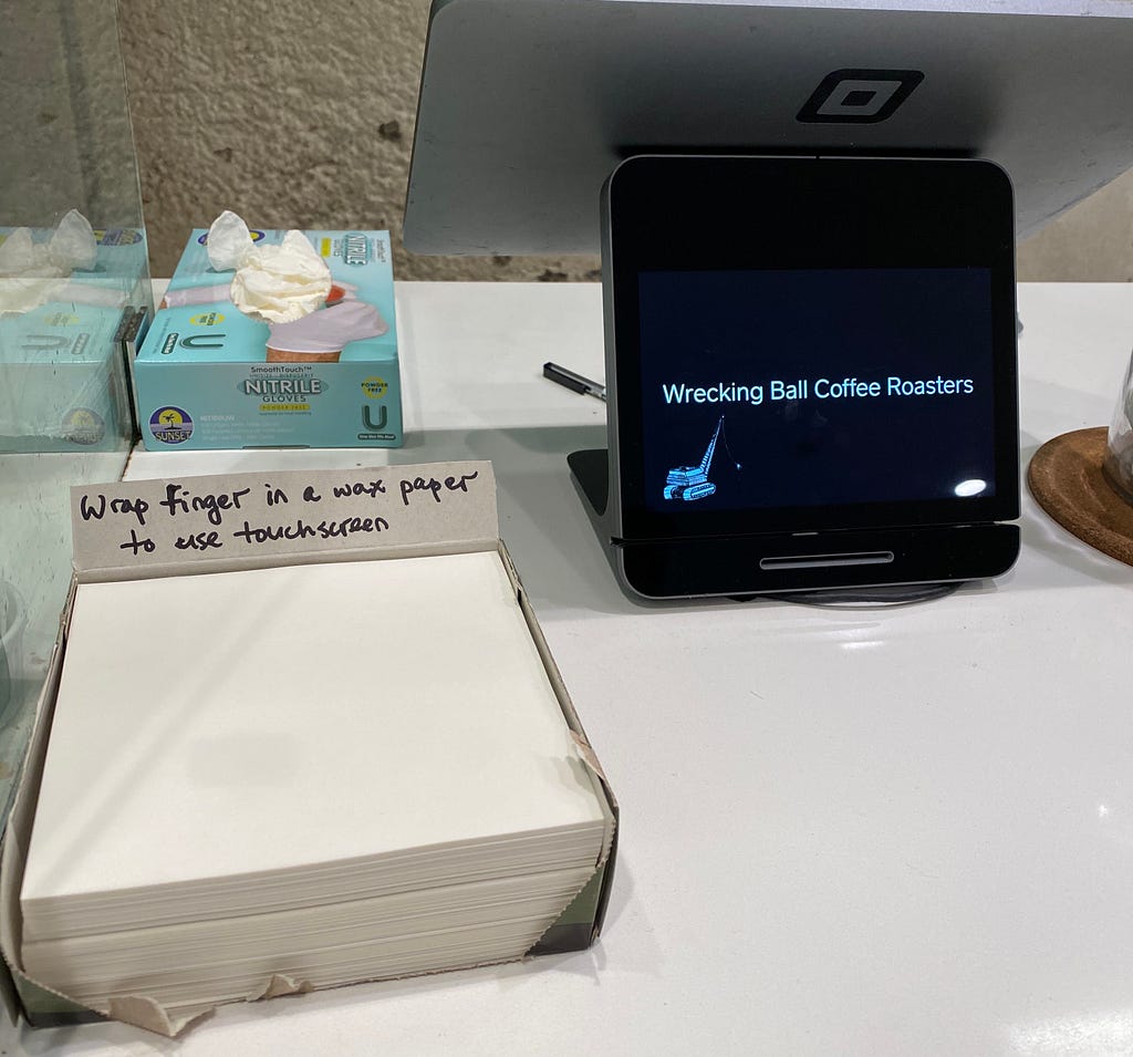 A stack of papers next to a cash register. There is a sign that reads “wrap finger in a wax paper to use touchscreen.”