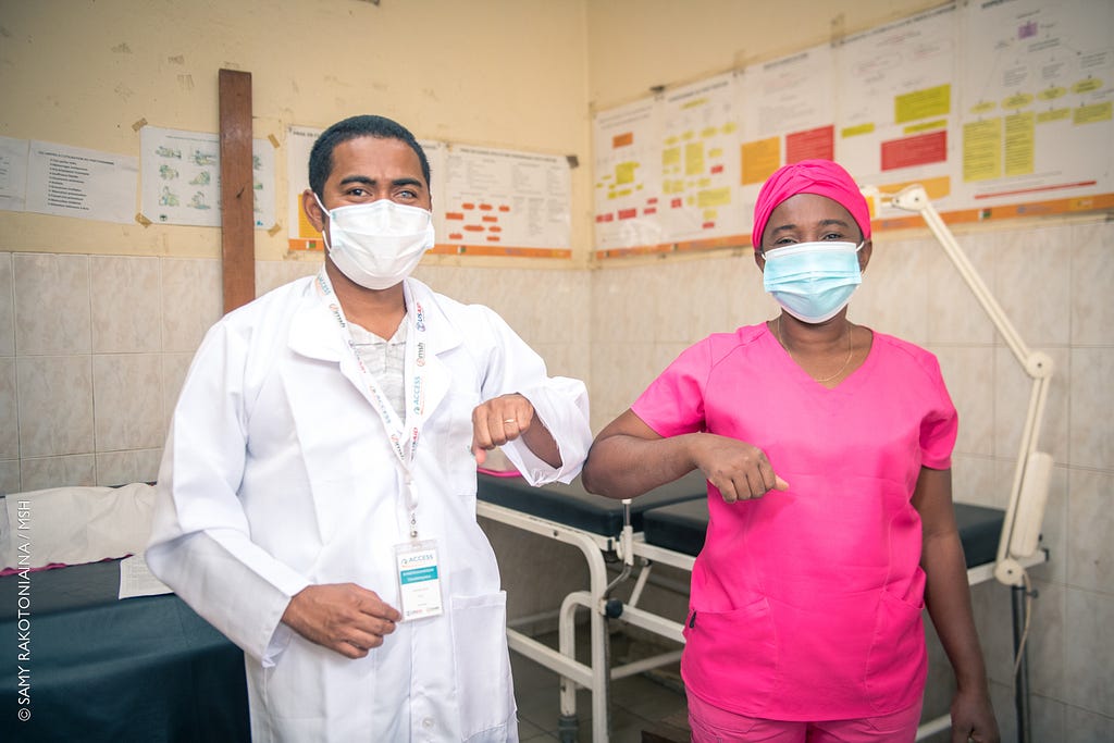Malagasy midwife Anasthasie, dressed in pink scrubs and headscarf, bumps elbows in solidarity with a health care trainer, who taught her modern infant resuscitation techniques.