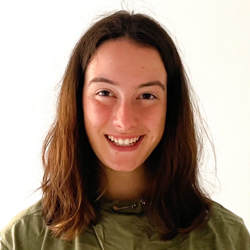 Photo of Sophia, a white woman, from the chest up. She is looking at the camera with an open lipped smile. She is wearing no makeup, a green and gold necklace and a green shirt. Her hair is naturally brushed and parted in the middle.