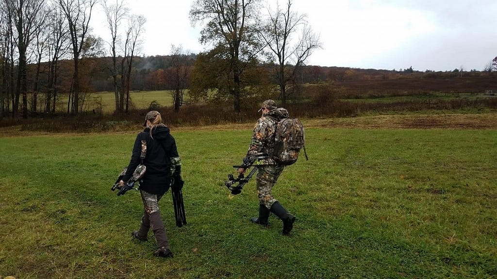 A man and a woman dressed in camo carry hunting gear across a field.