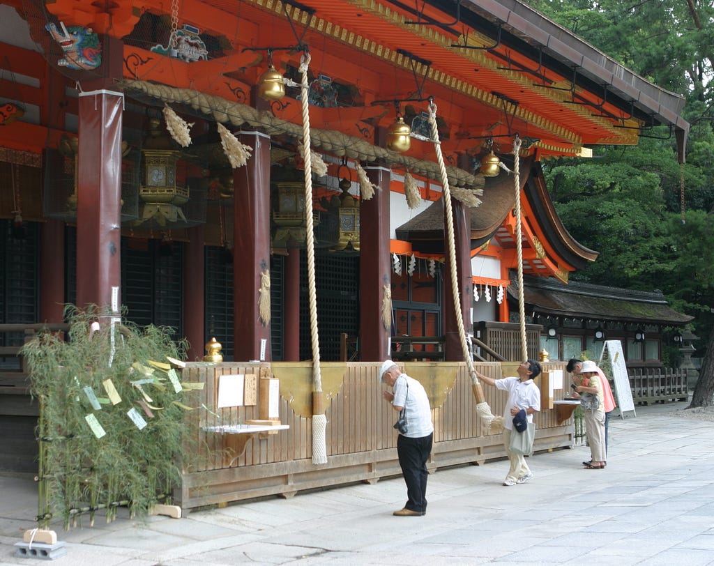 Four people stand before a Shinto shrine building. Long bellropes lead up to large bells hanging from the roof of the building. One person is using a rope to ring the bell, the others are praying.