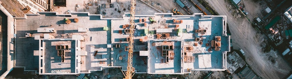 A top down view of a multi-story building under construction. A crane splits the middle of the image. The ground is covered with construction materials and eqiupment.