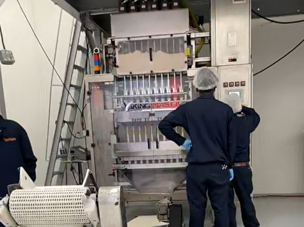 Inside factory with three workers wearing hairnets and blue factory uniforms, facing away from the camera.