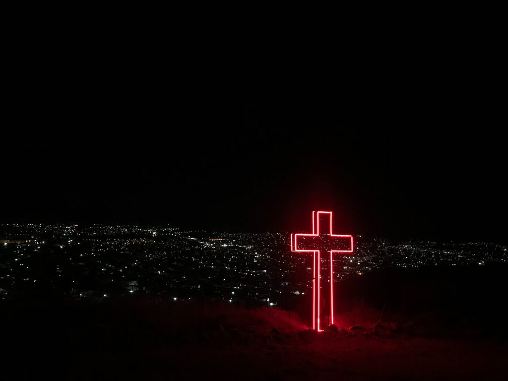 A red neon cross shines in the night over city lights in the distance