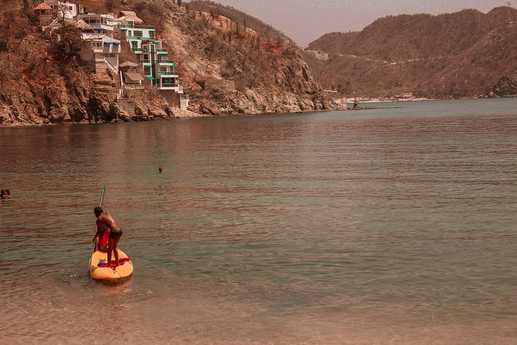 praia de ‘taganga’, entre montanhas o mar. a direita em um bote amarelo menino de pele negra remando em frente a risort.