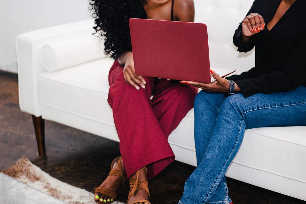 Two Black women sitting on a couch looking at a computer. The image shows only their legs and torsos.