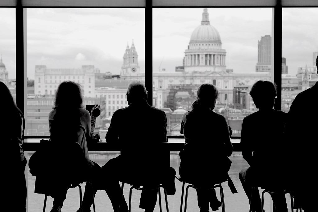 You can see four people from behind, sitting on bar stools and looking at the skyline of a city.