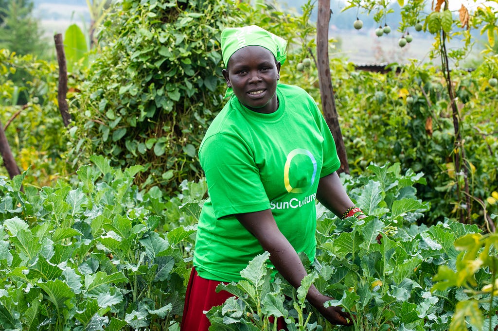 Rachael is centered in the photograph, smiling at the camera. She is surrounded by lush vegetation, including fruit trees and vegetable crops, with mountains visible far in the background.