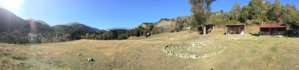 Green, tree-covered mountains below a blue sky. Two cabins on the right of the foreground, a stone labyrinth to their left.