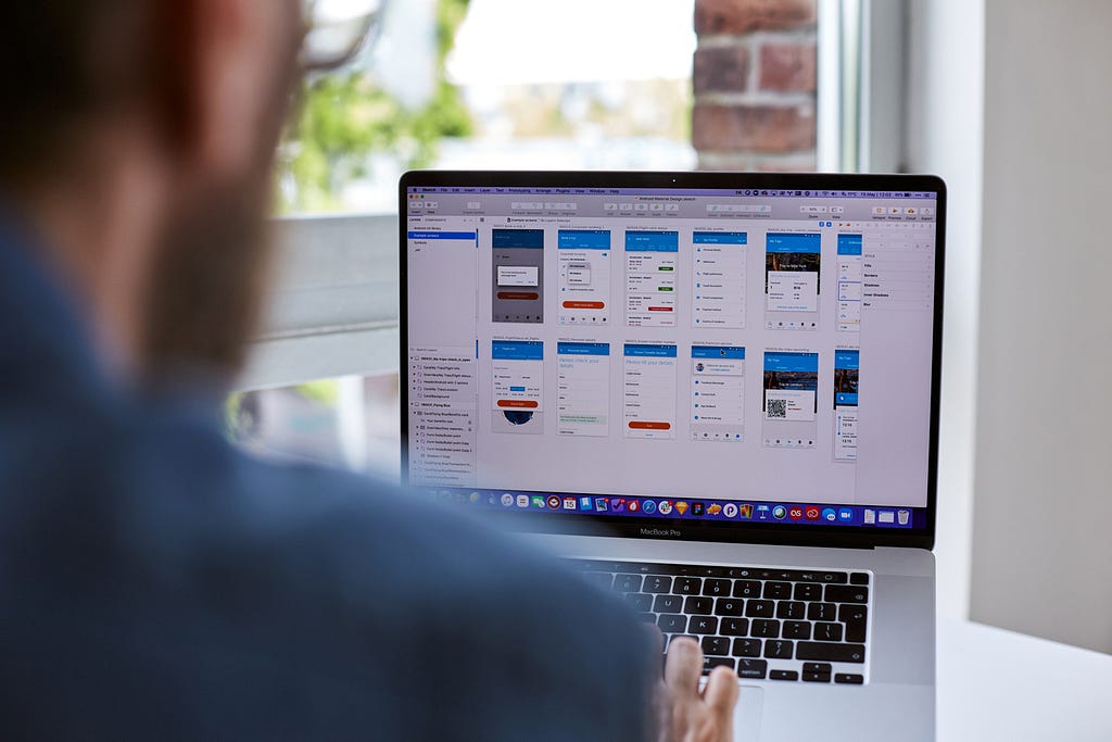 Dean sat in front of a computer, on screen are UI designs for the KLM app.