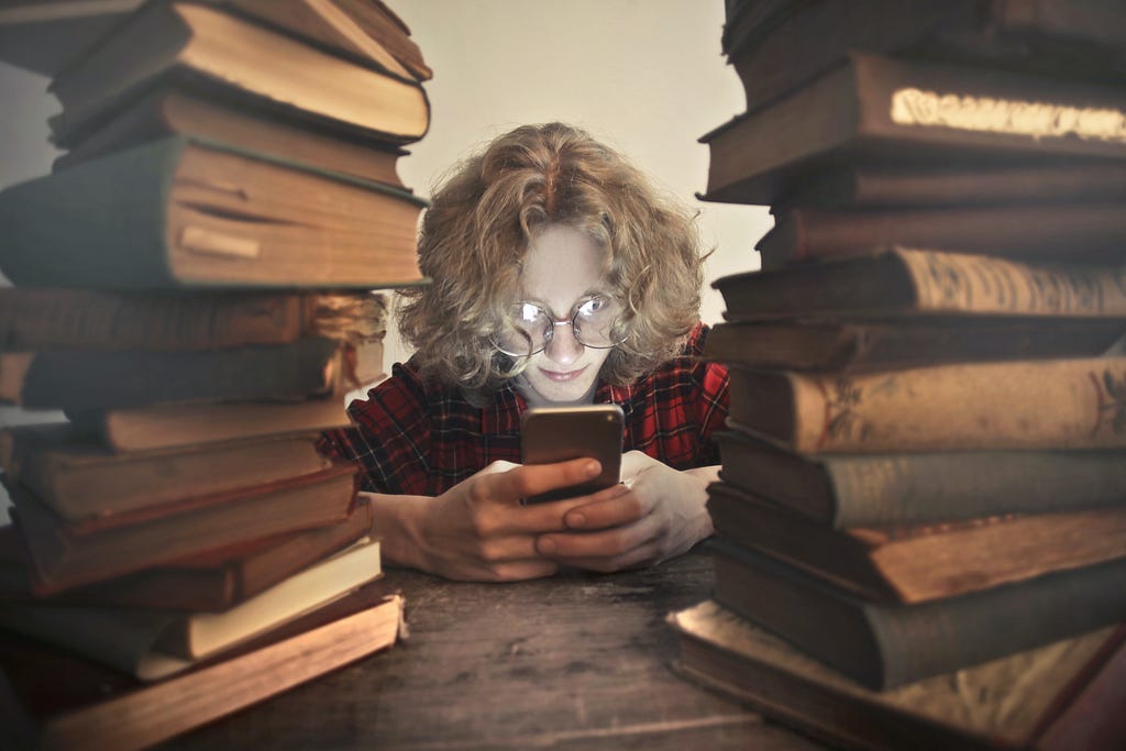 A person sitting between a stack of books, looking at their phone.