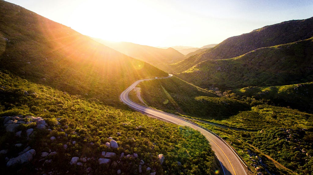 A photo of a road winding through verdant mountains towards the bright sun, image by Matt Howard