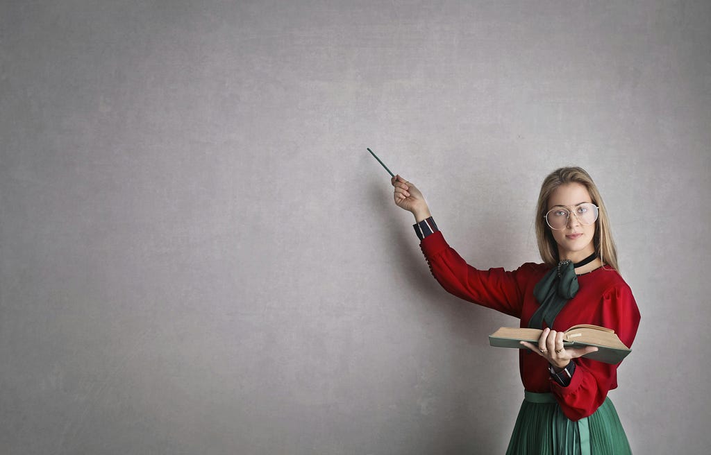photo entitled “Woman in Discussing A Lesson Plan.” she is dressed conservatively, holding a large open book in her left hand. she is holding a pointer in her right hand, which gestures at what appears to be a large blackboard where all the previous writings/markings have been erased.