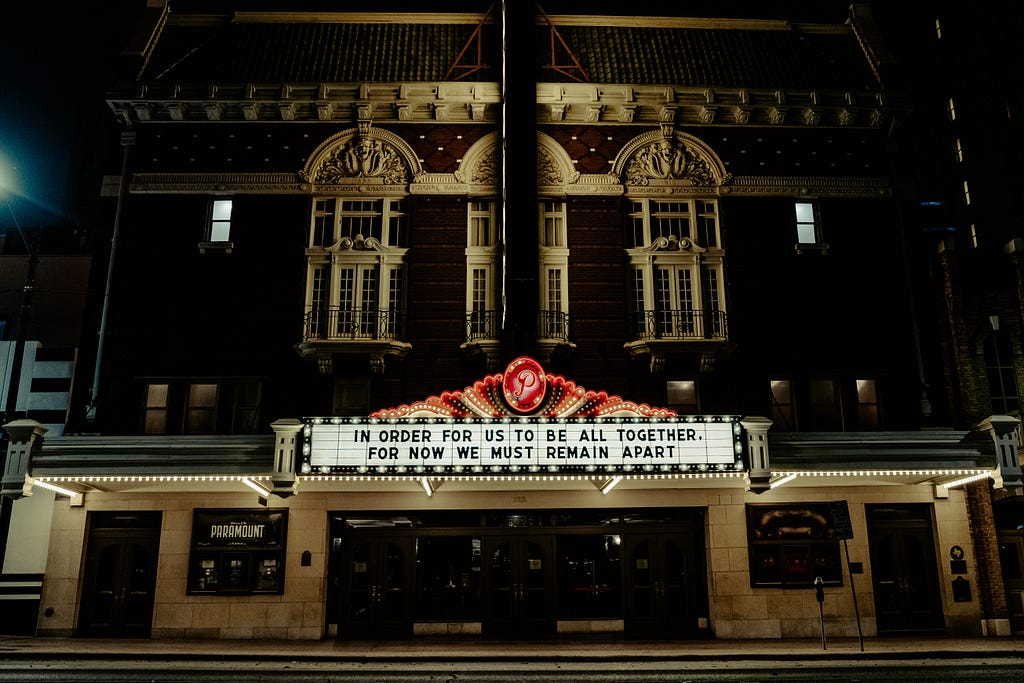 The front of the Paramount Theatre House (Austin, TX), with a marquis lit up in red, white, and black. The letters on the marquis read: “In order for us to be all together, for now we must remain apart.”