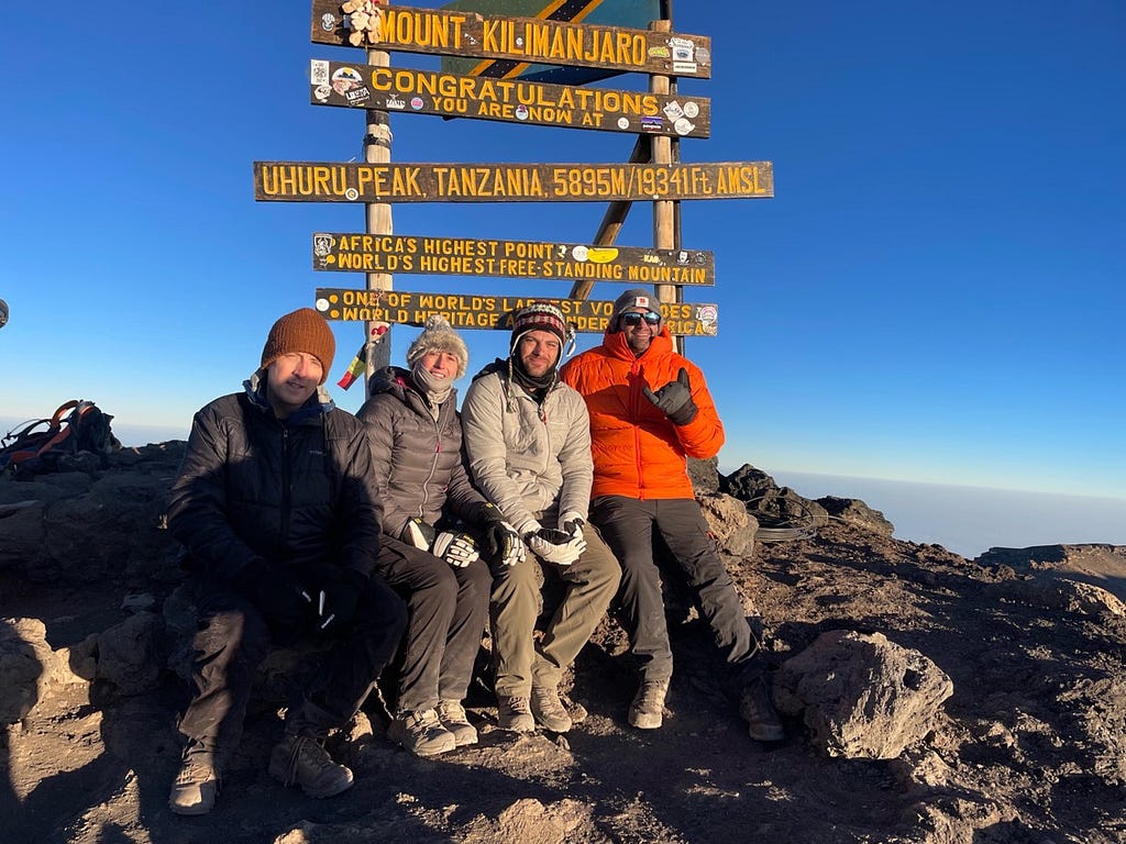 Author pictured with climbing mates, seated on boulders below the summit sign under blue sky at the top of Uhuru Peak on Mount Kilimanjaro.