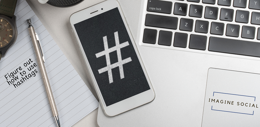 Hashtag symbol on a phone with a laptop and a piece of paper that says figure out how to use hashtags